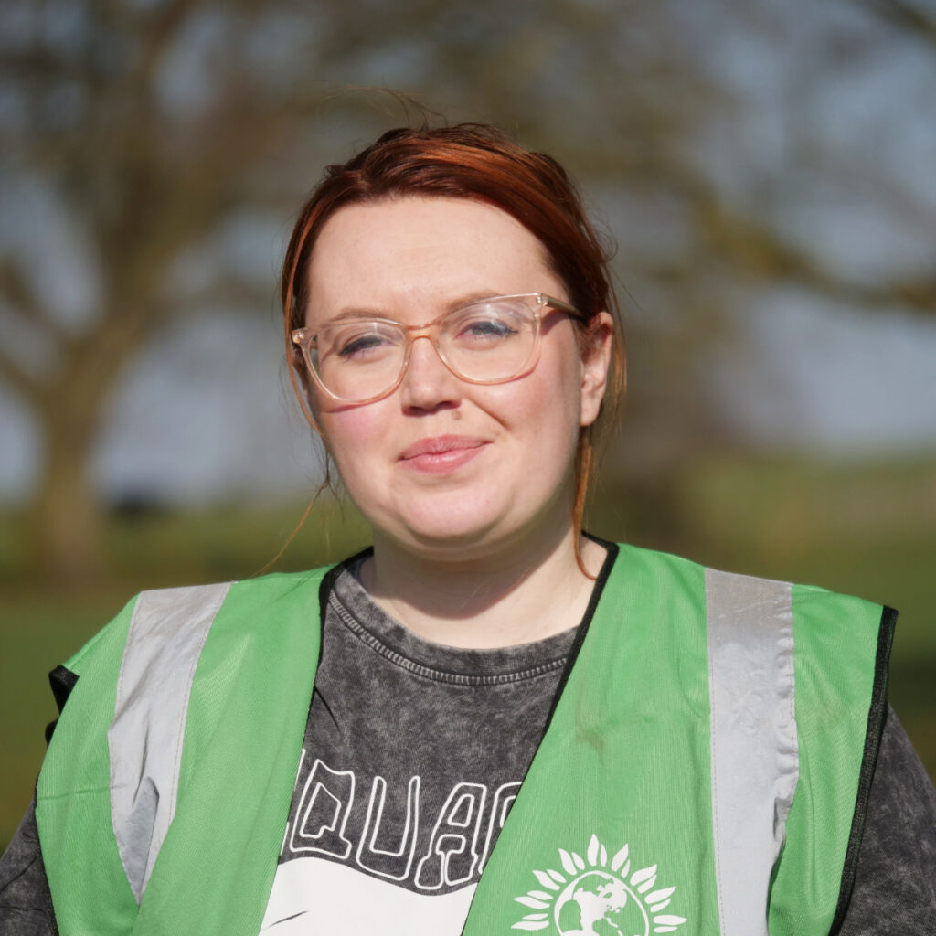 Headshot of Sarah Baumann standing in a field wearing a Green Party high-vis jacket